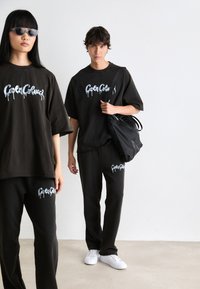 To modeller i matchende sorte sweatsets med hvite graffiti-stil logoer; en bærer en svart bag, mens den andre har på seg solbriller.