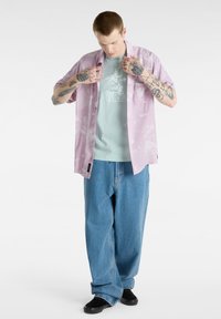 Camisa de manga corta de color rosa claro con un patrón floral sobre una camiseta gráfica verde menta, combinada con unos vaqueros de denim azul sueltos y zapatos negros.