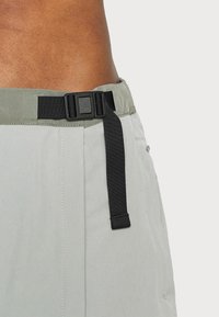 Ljusgrå shorts med ett brett, grönt midjeband som har en svart justerbar spänne och ett svart rem. Slät tygtextur med synlig sömnad.