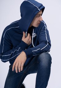 Marineblå zip-up hoodie med hvide striber, der har en hætte og et logo. Bæres med mørk denim jeans. Glat tekstur.