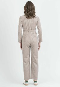 Mono de pana beige con mangas largas, cintura ajustada y bolsillos traseros; incluye un cinturón y se combina con zapatillas blancas y verdes.