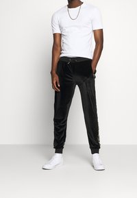 Eine Person steht selbstbewusst da, trägt ein weißes T-Shirt, schwarze Samtjogginghosen mit Seitenbranding und weiße Sneakers, mit den Händen in den Taschen.