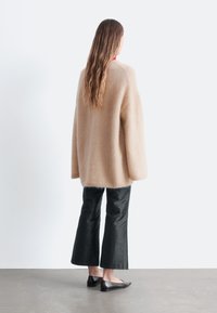 Übergroßer beiger Wollpullover, weite schwarze Hose und flache schwarze Schuhe, von hinten vor einem minimalistischen Hintergrund gesehen.