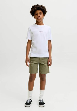Garçon debout, bras détendus, portant un t-shirt blanc, un short vert olive, des chaussettes blanches et des baskets noires sur un fond clair uni.