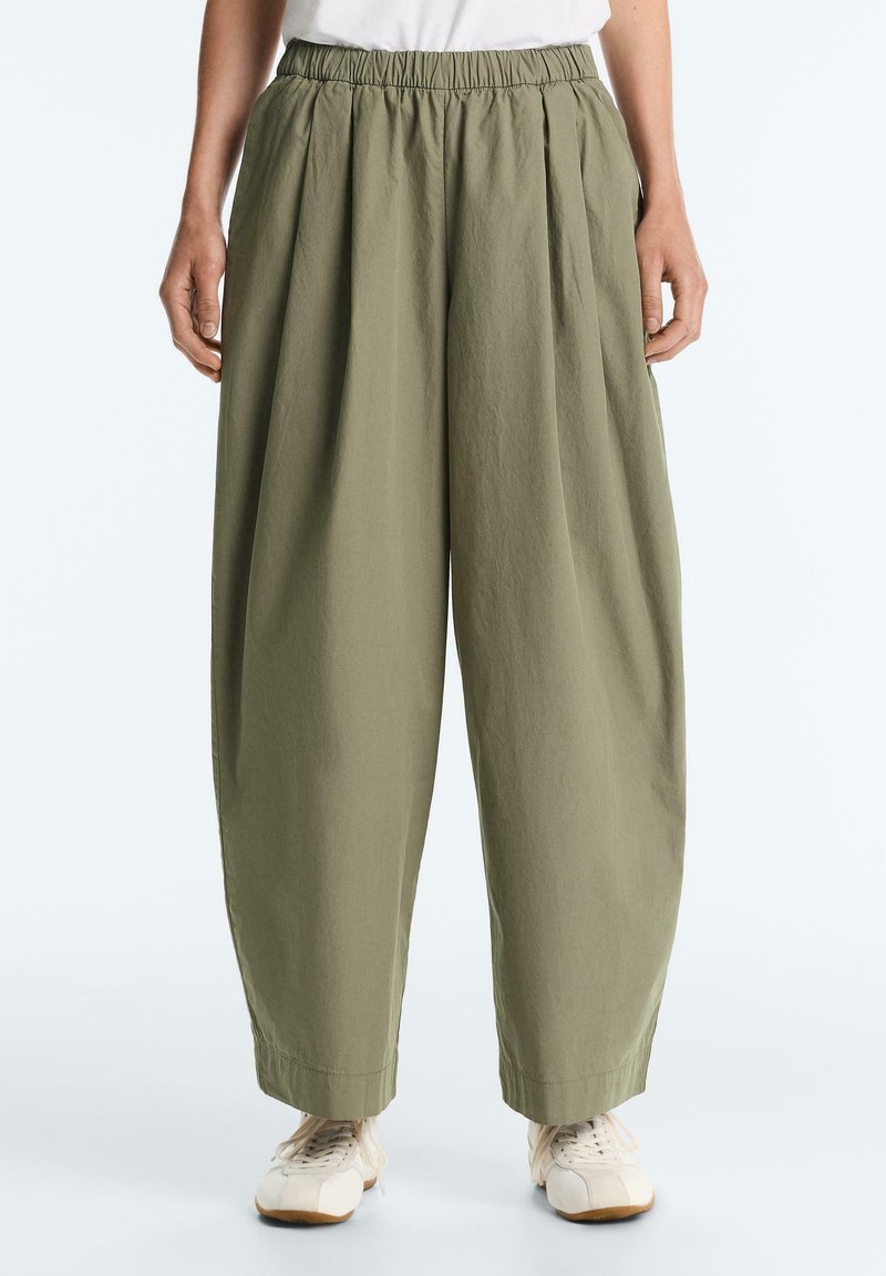 Person trägt locker sitzende, olivgrüne Hose mit elastischem Bund und weißen Turnschuhen, steht mit entspannten Händen seitlich am Körper.