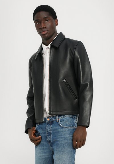 Chaqueta de cuero negra con cuello, cierre de cremallera y bolsillos laterales. Llevada sobre una camisa de rayas clara y combinada con jeans azules.