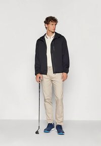 Νεαρός άνδρας ντυμένος με μαύρο μπουφάν, μπεζ παντελόνι και μπλε αθλητικά παπούτσια, κρατώντας ένα μπαστούνι γκολφ, στέκεται σε έναν απλό φωτεινό φόντο.