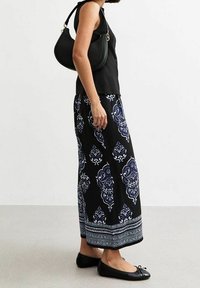 Černý top bez rukávů, dlouhá sukně s modro-bílým vzorem, ploché černé boty a černá zakřivená kabelka se zlatými kovovými detaily.