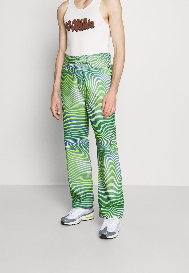 Zelené a modré kalhoty s vlnitým vzorem, vyrobené z hladké látky. V kombinaci s bílými a šedými sportovními botami.