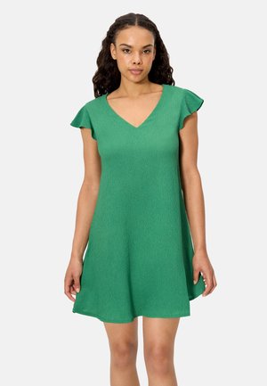 Mujer con un vestido corto verde de textura, con mangas flotantes y escote en V, posando frente a un fondo claro y liso.