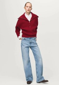 Roter, gerippter Pullover mit hohem Kragen und halbem Reißverschluss, kombiniert mit hellblauen, weit geschnittenen Jeans. Das Outfit wird mit einem weißen Hemd darunter ergänzt.