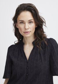 Blusa nera con colletto alla cinese, maniche corte a sbuffo e ricami decorativi con dettagli di bottoni sul davanti.