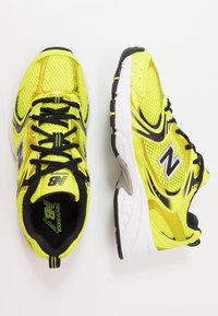 Ljusgula löparskor med ovandel i mesh, svarta accenter och en vadderad vit sula. Har svarta snören och en logotyp på sidan.