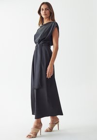 Black, satin gown with a one-shoulder design and tied waist. Smooth texture, floor-length, styled with beige high-heeled sandals.