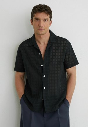 Mladý muž s krátkymi hnedými vlasmi, oblečený v čiernej krátkorukávovej košeli s gombíkmi a mriežkovým vzorom, stojaci pred bielou stenou.