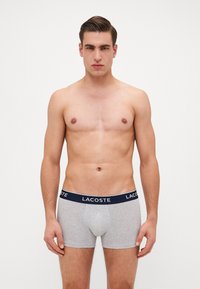 Boxer shorts de algodón gris con una cinturilla elástica en azul marino que presenta el logo en blanco de "LACOSTE". Textura suave y diseño ajustado.