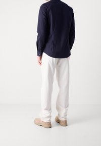 Marineblauw overhemd met lange mouwen en een button-down kraag, gecombineerd met loszittende witte broeken en lichtbeige schoenen. Vlakke textuur, minimalistisch ontwerp.