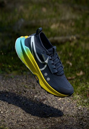 Mørkeblå løbesko med hvid swoosh og sål i gul-til-blå gradient, svævende over grussti i udendørs omgivelser.