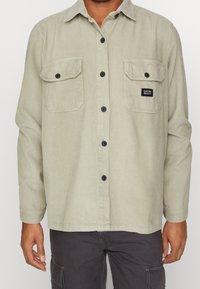 Jasnozielona sztruksowa koszula z długim rękawem, dwoma kieszeniami na piersi, czarnymi guzikami i małą metką na lewej kieszeni. Teksturowana tkanina.