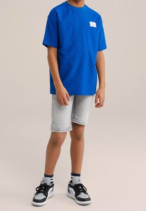 Chlapec v královsky modrém tričku s krátkým rukávem, světle šedých džínových šortkách, černobílých teniskách a tmavě modrých ponožkách na jednobarevném béžovém pozadí.