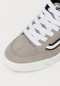 Bout de sneaker en daim gris avec lacets blancs, bande latérale noire et blanche, et semelle en caoutchouc blanc texturé sur un fond clair.