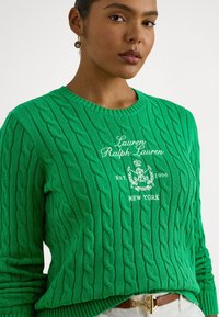 Zielony sweter w sploty z prążkowanym dekoltem, białym haftowanym logo i fakturowanym wzorem. Noszony z jasnym paskiem.