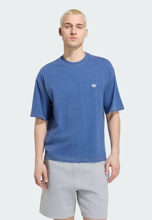 Ung mand med kort blondt hår iført en blå Adidas t-shirt og lysegrå shorts, stående mod en ensfarvet lys baggrund.
