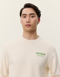 Krém színű pulóver zöld logóval a mellrészen, kerek nyakkivágással és bordázott mandzsettával. Simább anyag, laza szabás.