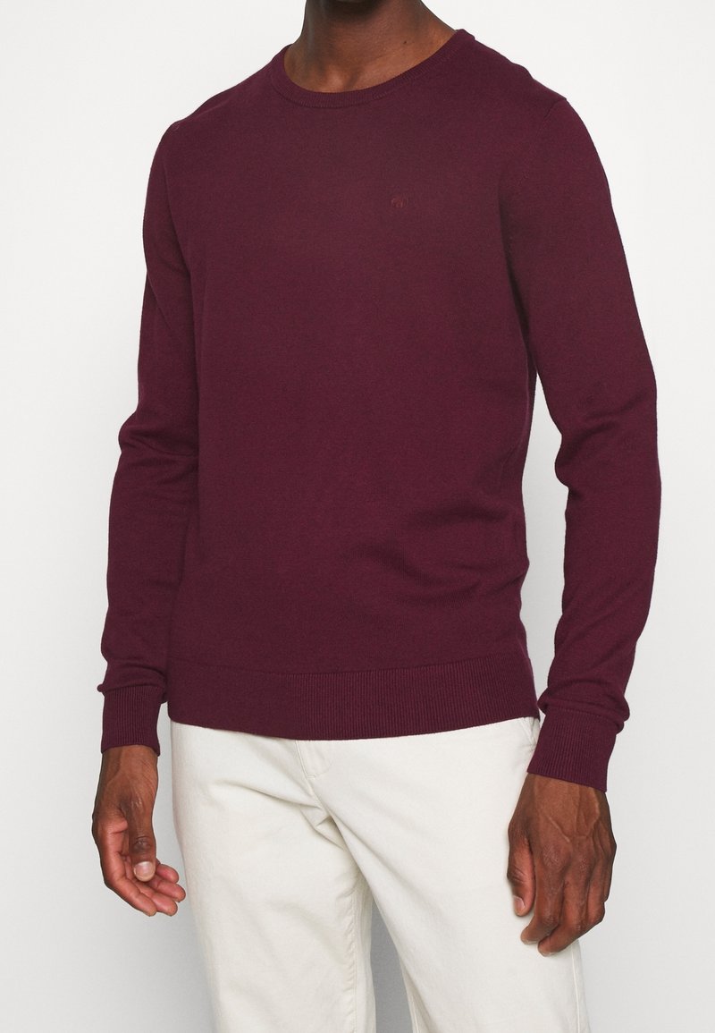 Burgundy knitted sweater with a crew neckline, long sleeves, and ribbed cuffs and hem. Subtle logo detail on the chest.