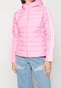 Vaaleanpunainen toppatakki, jossa on vaakasuora tikattu kuvio, vetoketju edessä ja valkoiset pystysuorat raidat hihoissa. Takissa on huppu ja sivutaskut.