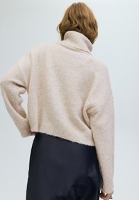 Világos rózsaszín, túlméretezett pulóver magas gallérral, texturált kötött mintával és leejtett vállak. Fekete, csipkedíszítéses szoknya fölött viselve.