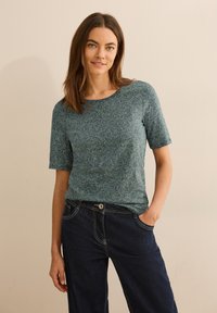 Tyrkysové tričko s krátkým rukávem, geometrickým vzorem, kulatým výstřihem a měkkou texturou, v kombinaci s tmavě modrými širokými kalhotami.