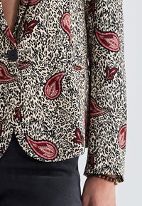 Kavaj i krämfärgad med ett svart och rött paisleymönster, med slagkrage, en knappstängning och två framfickor.