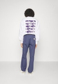 Sudadera blanca con texto morado en la parte posterior, combinada con pantalones azul marino. La sudadera cuenta con cuello redondo y puños acanalados.