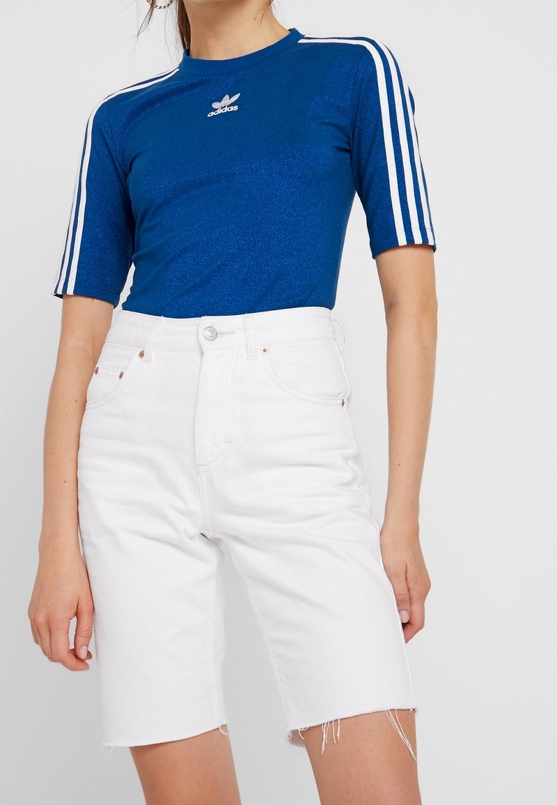 Modrý top s krátkým rukávem s lesklou texturou a bílým logem Adidas, doplněný o bílé džíny šortky s roztrhaným lemem.