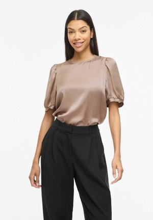 Kobieta o długich ciemnych włosach, ubrana w błyszczącą beżową bluzkę z bufiastymi rękawami, wpuszczoną do wysokich, czarnych plisowanych spodni, stojąca i uśmiechająca się.