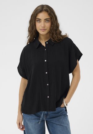 Mladá žena s vlnitými hnedými vlasmi, oblečená v voľnej čiernej košeli s krátkym rukávom a modrých džínsach, pózujúca s jednou rukou v vrecku.