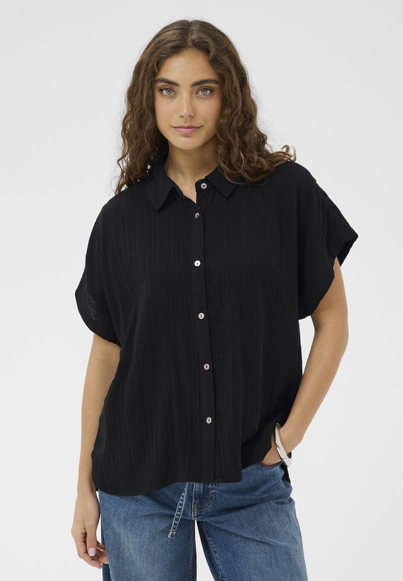 Noor pruunide laineliste juustega naine, kes kannab lahtist musta lühikeste varrukatega nööbitavat särk ja siniseid teksapükse, poseerides ühe käega taskus.