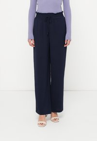 Γυναίκα που φοράει ναυτικές παντελόνες με φαρδύ τελείωμα και ελαστική μέση με κορδόνι, σε συνδυασμό με μπεζ ανοιχτές τις μύτες γόβες και μωβ μπλούζα με μακριά μανίκια.