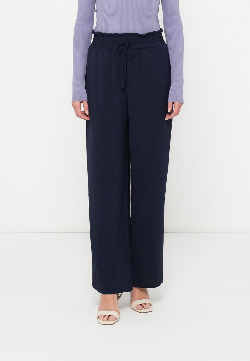 Γυναίκα που φοράει ναυτικές παντελόνες με φαρδύ τελείωμα και ελαστική μέση με κορδόνι, σε συνδυασμό με μπεζ ανοιχτές τις μύτες γόβες και μωβ μπλούζα με μακριά μανίκια.