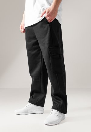 Persona che indossa pantaloni cargo neri con tasche laterali e sneakers bianche, in piedi con una mano in tasca su sfondo semplice.