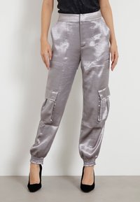 Mujer vestida con pantalones cargo plateados brillantes con puños elásticos y tacones altos negros, de pie contra un fondo neutro.