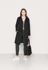 Cappotto trench nero doppiopetto con vita cintrata, indossato sopra un maglione verde e una camicia a righe bianche. Abbinato a pantaloni verdi e sneakers beige.