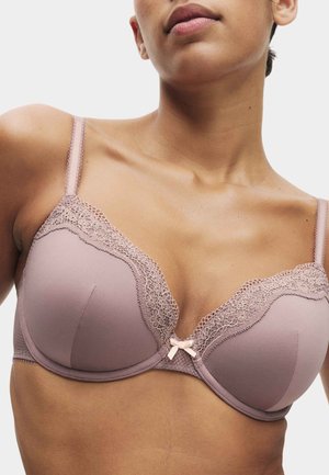 Kvinde iført en lys mauve bh med blondetrim og en lille sløjfe i midten, vist fra skuldre til øverste del af brystet mod en ensfarvet baggrund.