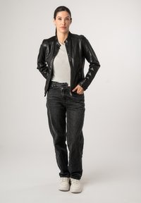 Schwarze Lederjacke mit Kragen, Reißverschluss vorne und strukturiertem Finish. Getragen über einem hellgrauen Oberteil und kombiniert mit dunklen Jeans und weißen Sneakers.