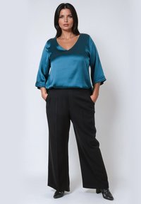 Woman with dark hair wearing a teal satin V-neck blouse, black wide-leg trousers, and black pointed boots, standing with hands in pockets.
