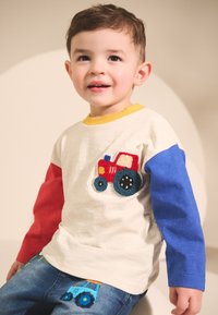 Maglietta bianca con maniche rosse e blu, con un trattore rosso ricamato. Indossata con jeans blu decorati con un disegno di trattore.