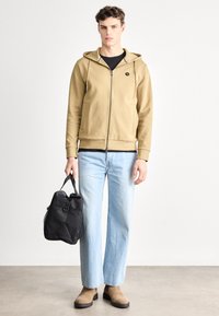 Khaki mikina na zips s šnúrkami, pod ňou čierne tričko, svetlomodré džínsy, béžové členkové čižmy a čierna taška. Hladká látka, voľný strih.