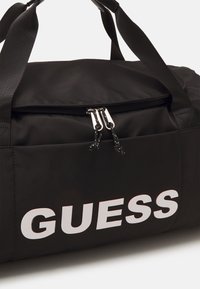 Borsone nero in nylon con doppia cerniera argentata e grande logo "GUESS" in bianco sul lato. Dotato di manici piatti e robusti.