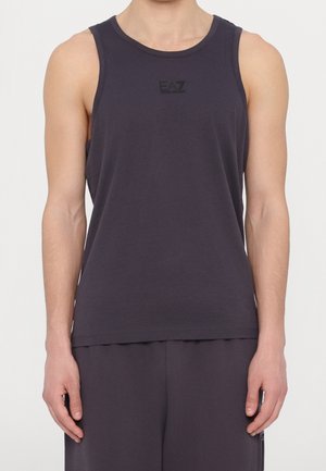 Mężczyzna w bezrękawniku w ciemnym granatowym kolorze z logo EA7 Emporio Armani oraz dopasowanych ciemnych spodniach, stojący na tle jednolitej białej ściany.